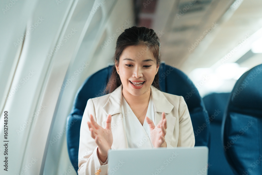 Asian businesswoman gesturing and smiling during video conference on laptop while sitting in airplane seat, concept of remote work and business travel Asian businesswoman gesturing and smiling during video conference on laptop while sitting in airplane seat, concept of remote work and business travel