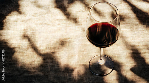 A glass of red wine sits on a textured surface with shadows.