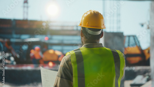 Construction manager supervising building site holding blueprints wearing safety vest and helmet
