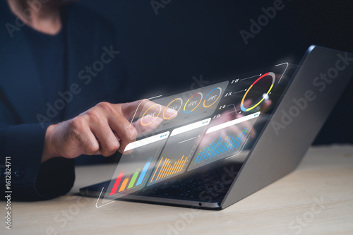 A person interacts with a futuristic transparent touchscreen displaying colorful charts and graphs above a laptop.