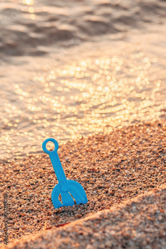 Fototapeta Naklejka Na Ścianę i Meble -  Blue plastic shovel stuck in pebbly beach sand with golden light reflecting off the gentle sea waves.