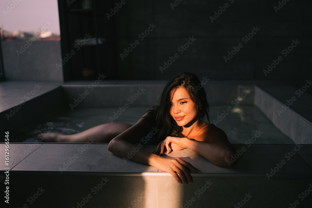 Obraz premium A woman relaxes in a sunlit bath, the water glistening as she leans on the edge, embraced by warm light and serene ambiance of a luxurious setting.