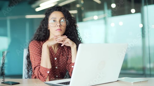 Busy businesswoman thinking about the task while working on laptop sitting at workplace in a modern business office. Thoughtful young female employee deals with project, thinks about solving a problem