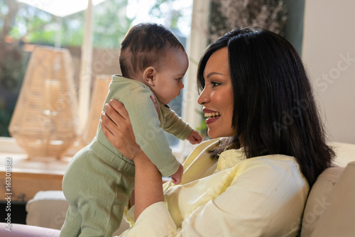 Asian mother lifting daughter wearing pale green onesie on beige sofa near wooden console