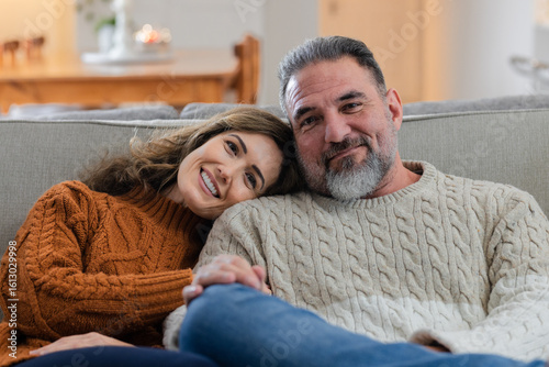 Senior couple sitting on light-gray sofa in cozy living room beside wooden sideboard and lit candle