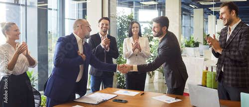 Bilde på lerret Successful business team partners shaking hands across a table after signing a contract, surrounded by smiling colleagues clapping and celebrating in modern glass office environment