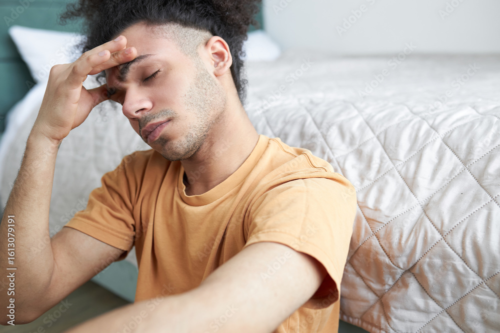 Obraz premium Young man in orange shirt holding his forehead with closed eyes, suffering migraine or headache, sitting on floor in his bedroom with closed eyes, feeling unwell after waking and getting up