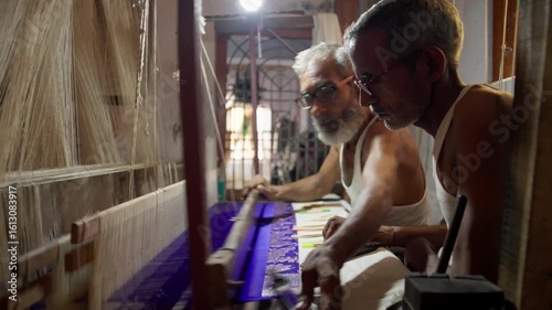 Close-up of two artisans weaving a Banarasi saree with gold thread on handloom in Varanasi, India. Focus on hands, intricate design, and traditional silk weaving process.
