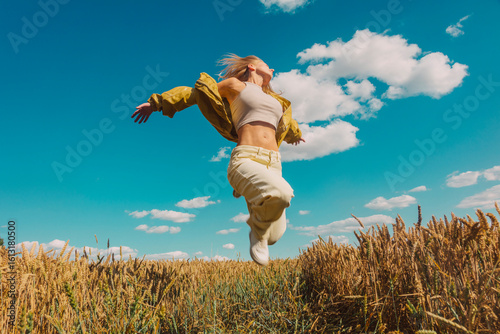 Stylish woman with blonde hair jumping in wheat field under blue sky