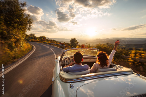 Photography Couple driving a vintage convertible car on a winding road at sunset with one person raising their hand joyfully