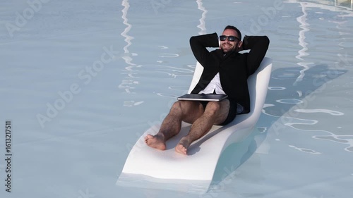Businessman relaxing on a lounge chair in a pool, wearing sunglasses and a suit, enjoying leisure time while holding a laptop, embodying work-life balance and tranquility