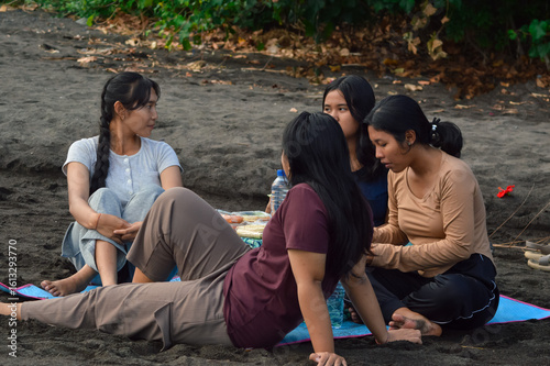 Four girls sit comfortably on a mat on the sandy beach, surrounded by trees and dry leaves, engaged in cheerful conversation while enjoying their light morning picnic.