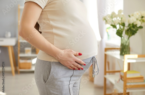 Cropped image of unrecognizable pregnant woman expecting mother touching belly, has big tummy, being on third trimester, standing against cozy home background. Pregnancy and expectations concept