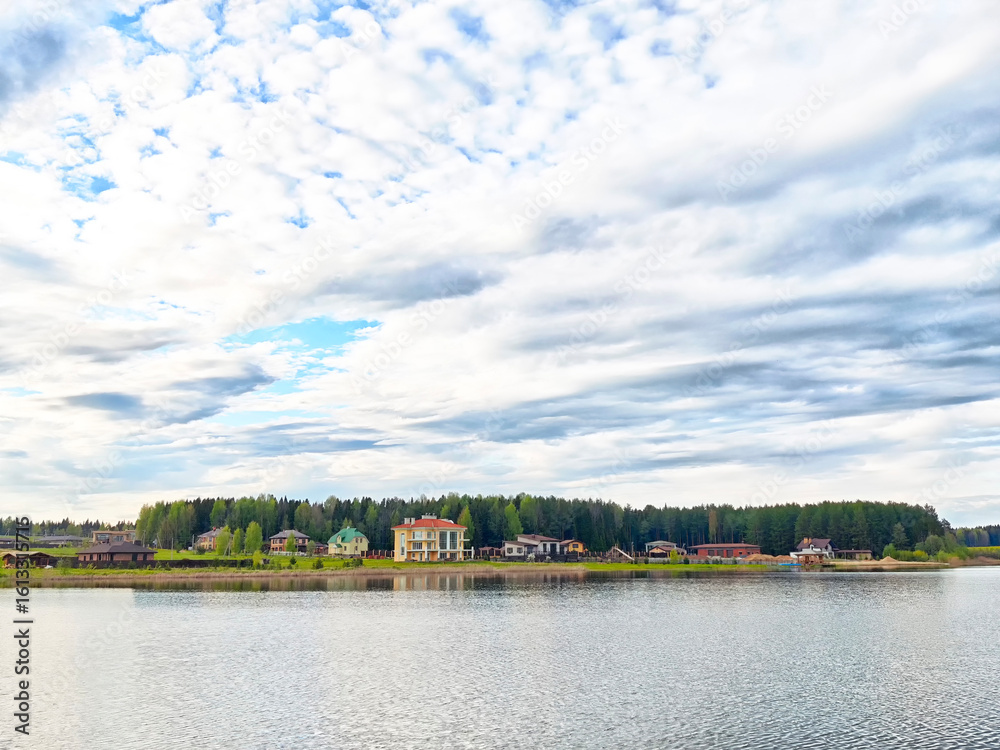 Fototapeta premium Calm lake view with colorful houses amid lush green trees under a cloudy sky in a serene rural setting