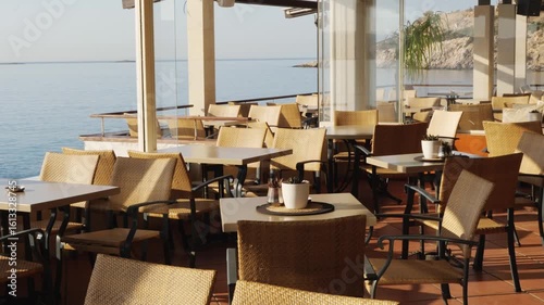 Wallpaper Mural Spacious restaurant in Varkiza, Greece, with wicker chairs, terracotta tile floor, white plant pots, and wide windows opening to clear sea and rocky shore. No people, natural sunlight Torontodigital.ca