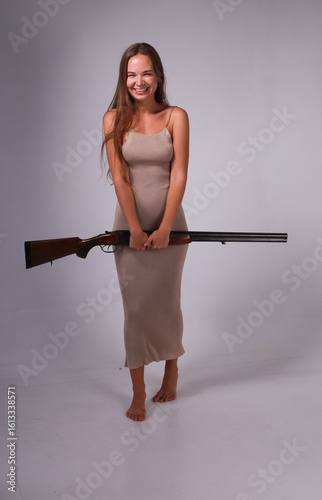 Slender barefoot young woman in long dress holding double barrelled vintage shotgun on grey background full length