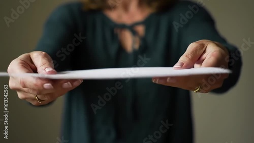 Person extends blank white paper towards viewer. Hands visible, wearing a green blouse against a muted background