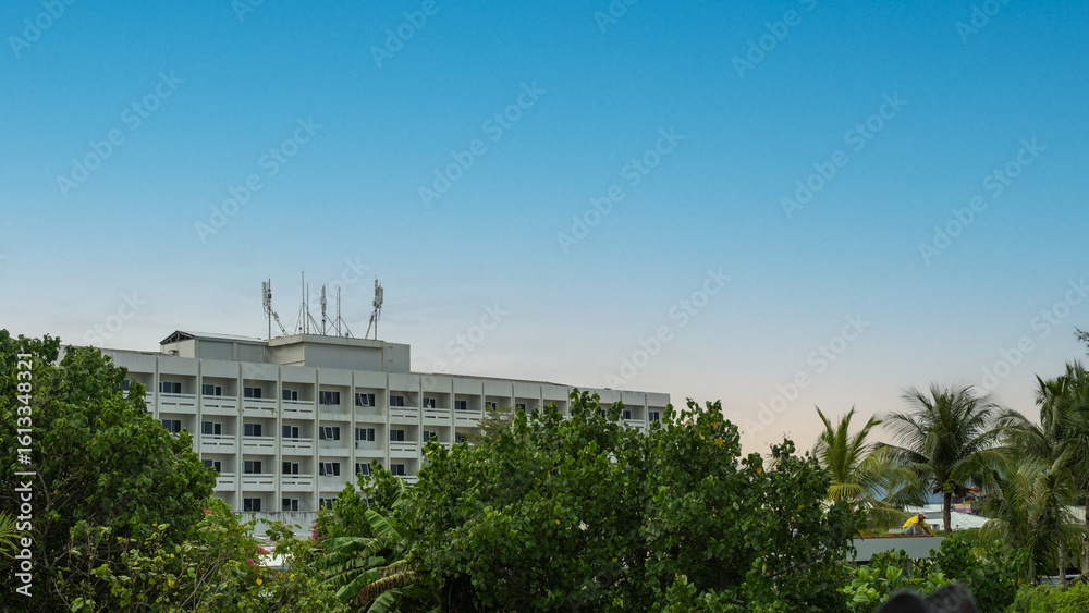 Fototapeta premium Tropical urban landscape with a mid-rise building, telecommunication antennas on the roof, and lush green trees under a clear blue sky at sunrise.