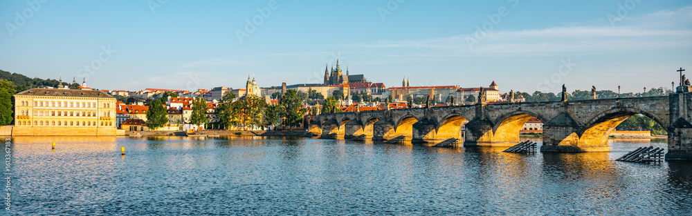Fototapeta premium Breathtaking panorama showcases Prague Castle rising above the Vltava River, with the iconic Charles Bridge spanning the calm waters, reflecting the morning light in a serene cityscape.