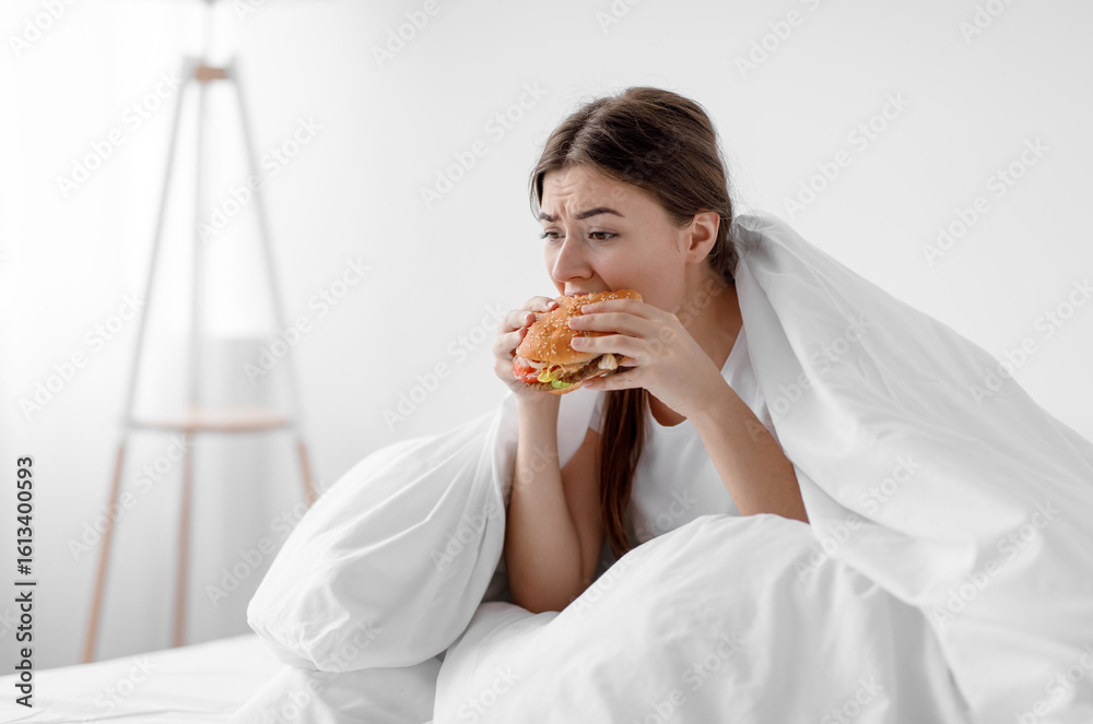 Starving upset hungry cute caucasian millennial woman sitting in bed at home and eating burger suffering from stress, depression, crisis and gluttony. Junk food, substitution, diet and too much meal Starving upset hungry cute caucasian millennial woman sitting in bed at home and eating burger suffering from stress, depression, crisis and gluttony. Junk food, substitution, diet and too much meal