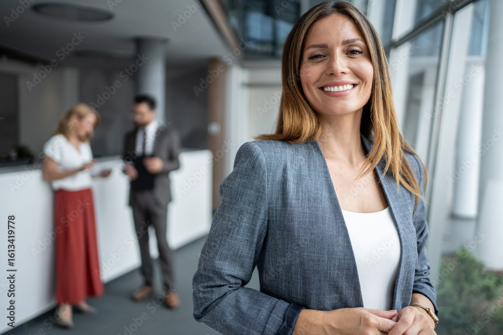 Fototapeta premium Smiling businesswoman standing in modern office with colleagues discussing in background