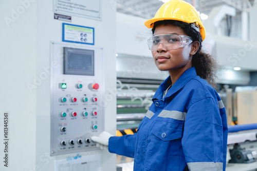 Portrait of Hispanic female technician in safety gear operating control panel at modern manufacturing plant, smiling confidently at camera.