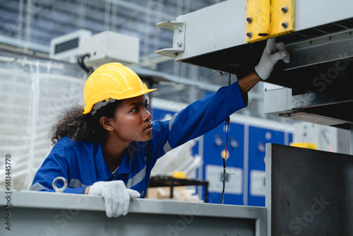Female industrial worker wearing a yellow helmet and blue uniform examines a cardboard processing machine in a modern paper box factory, showing precision and safety in her task.
