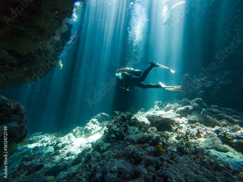 Landscape view of sunlight sunbeams dancing from the surface in underwater sea cave with rock walls and scuba diver exploring