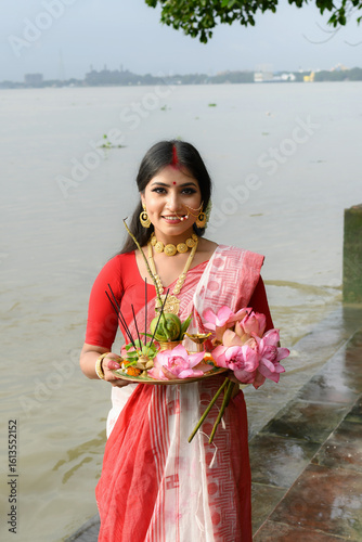Portrait of a beautiful young Indian woman in a traditional saree, wearing gold jewellery and bangles, holding a plate filled with religious offerings in front of the river Ganges