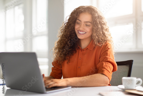 Obraz na płótnie A radiant young woman in an orange shirt works on her laptop with a beaming smil