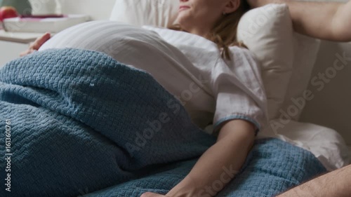 Tilting medium close-up of hands and face of Caucasian man visiting heavily pregnant wife in maternity hospital, stroking hair. Exhausted woman in white medical gown sleeping in bed