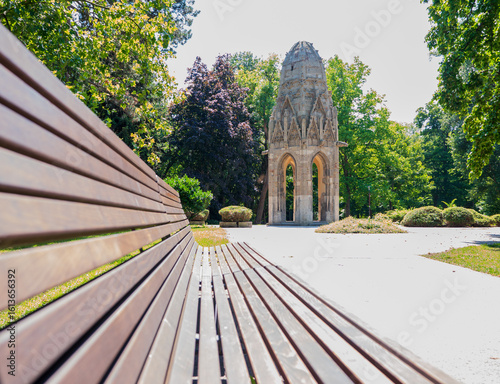 Photography Old gothic tower in Sad Janka Kráľa park in Bratislava, Slovakia, with wooden bench in foreground and green trees in the background on a sunny summer day