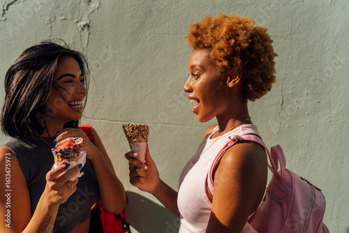 Two happy female friends with ice cream cones talking at a wall