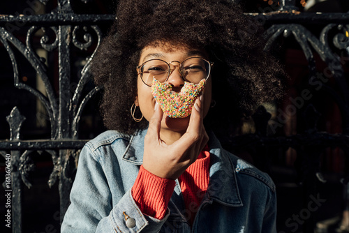 Close-up of young woman with curly hair eating donut against fence in city