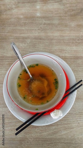 A close-up of a bowl of clear broth with chopped green onions, a spoon, and chopsticks.