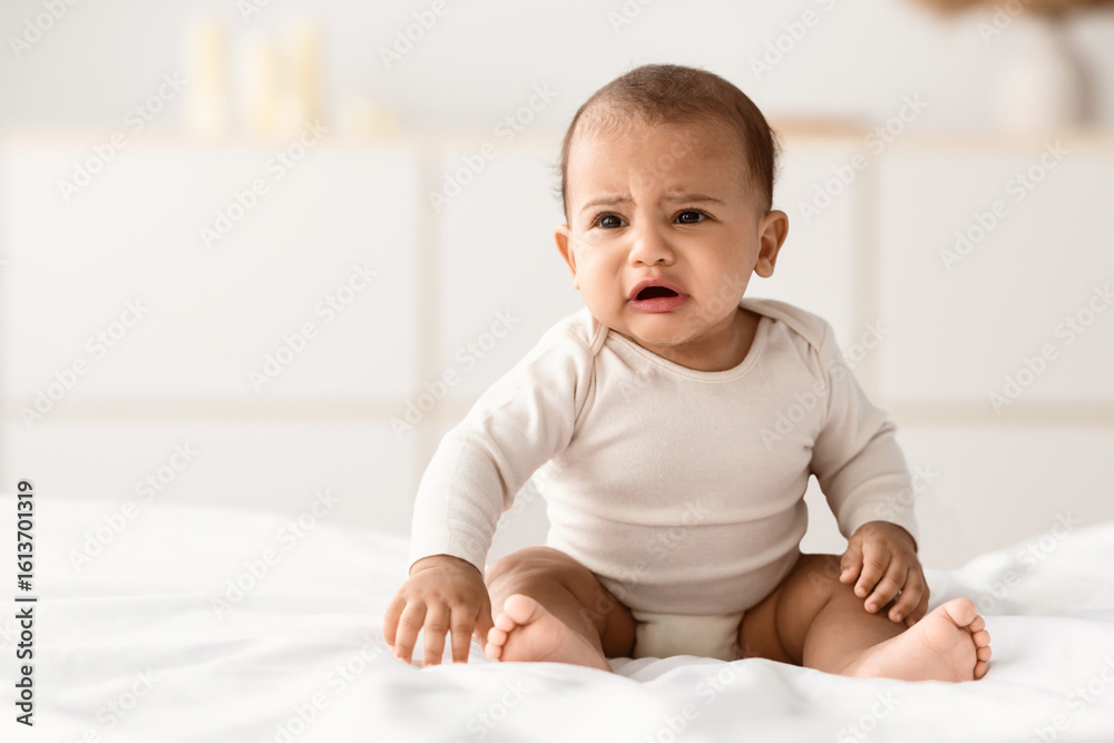 Φωτογραφία Portrait of sad upset African American baby crying, sitting on the bed in bedroom at home, wearing bodysuit