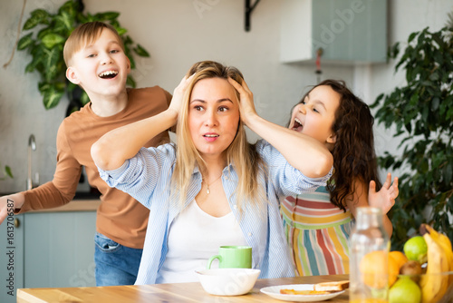 Young woman mom sitting on a kitchen table looking stressed suffers from headache because of noise of little children