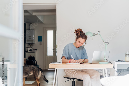 Female professional writing in book while sitting at table in home office
