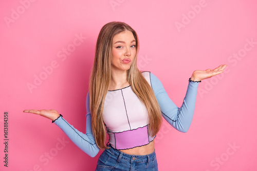 Charming young woman with long blonde hair posing in casual colorful top against pink background with playful expression