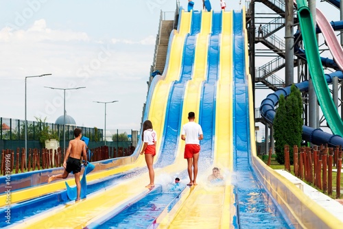 Children and adults are enjoying a blue and yellow water slide in Aqualand. Some people are waiting to slide down, while others are enjoying the cool water at the bottom of the slide on a sunny day