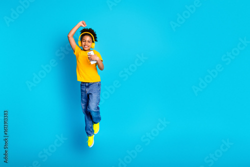 Cheerful young girl with headphones and phone jumping joyfully against a vibrant blue background