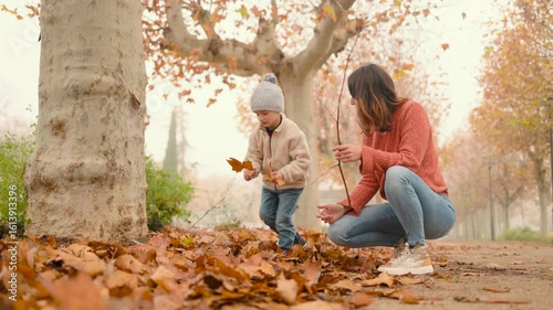 Happy mother and son are collecting fallen leaves in a park during a beautiful autumn day.