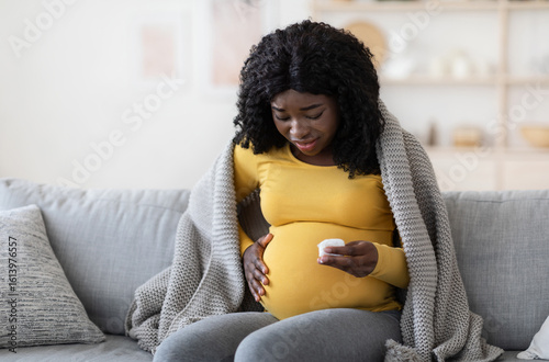 Pregnant black woman covered in blanket feeling sick, sitting on couch at home, holding napkin, worrying about her baby, empty space. Expecting african american lady having cold or flue