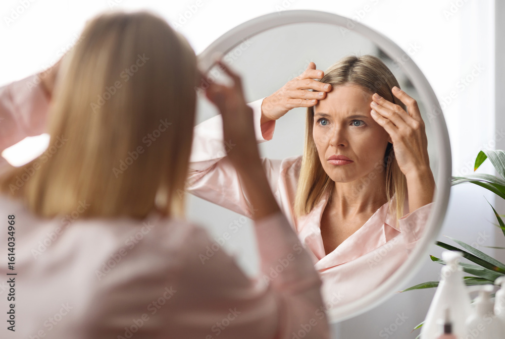 Naklejka premium Middle Aged Woman Looking In Mirror And Touching Wrinkles On Her Face, Upset Beautiful Mature Female Examining Fine Lines On Forehead, Suffering Skin Aging, Selective Focus On Reflection, Closeup