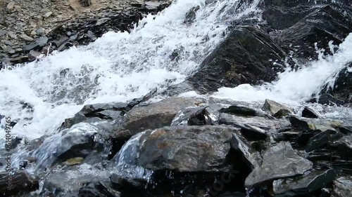 A tranquil mountain stream flows over dark, rugged rocks, creating a gentle cascade as sunlight glistens off the water's surface