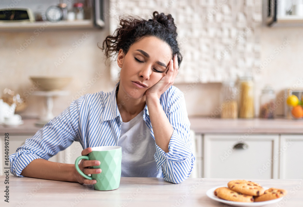 Obraz premium Sleepy young woman sitting at dining table in kitchen with closed eyes, holding cup while having breakfast. Early wake up, insomnia, lack of sleep