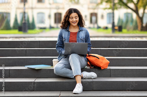 Cuadro en lienzo Joyful young indian woman student studying online, using laptop computer outdoors, sitting on stairs at college campus, doing homework, copy space