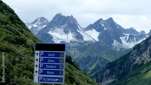 Snow-capped alpine peaks rise majestically above green valleys, with a roadside parking sign pointing toward Lech, Zürs, and Stuben in the Austrian Alps.