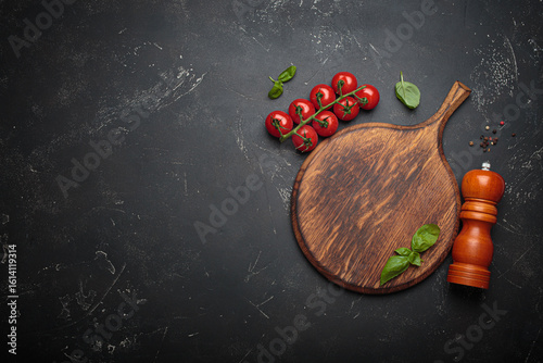 Fototapeta Naklejka Na Ścianę i Meble -  Cherry tomatoes, garlic and fresh basil leaves arranged on a rustic wooden cutting board beside a wooden pepper mill against a dark slate textured background, copy space