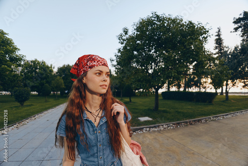 Lifestyle woman in modern boho-western denim street style wearing a red bandana, walking outdoors during golden hour with natural greenery and warm film color tones.